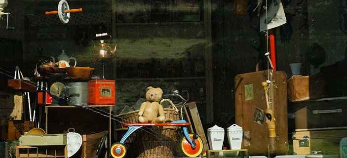 Schaufenster der Ladenstube im Antik- und Trödelladen Swen Homrighausen in Bad Berleburg-Wingeshausen mit Teddybär auf altem Dreirad, Vintage-Koffer, antiken Blechdosen und Sammlerstücken